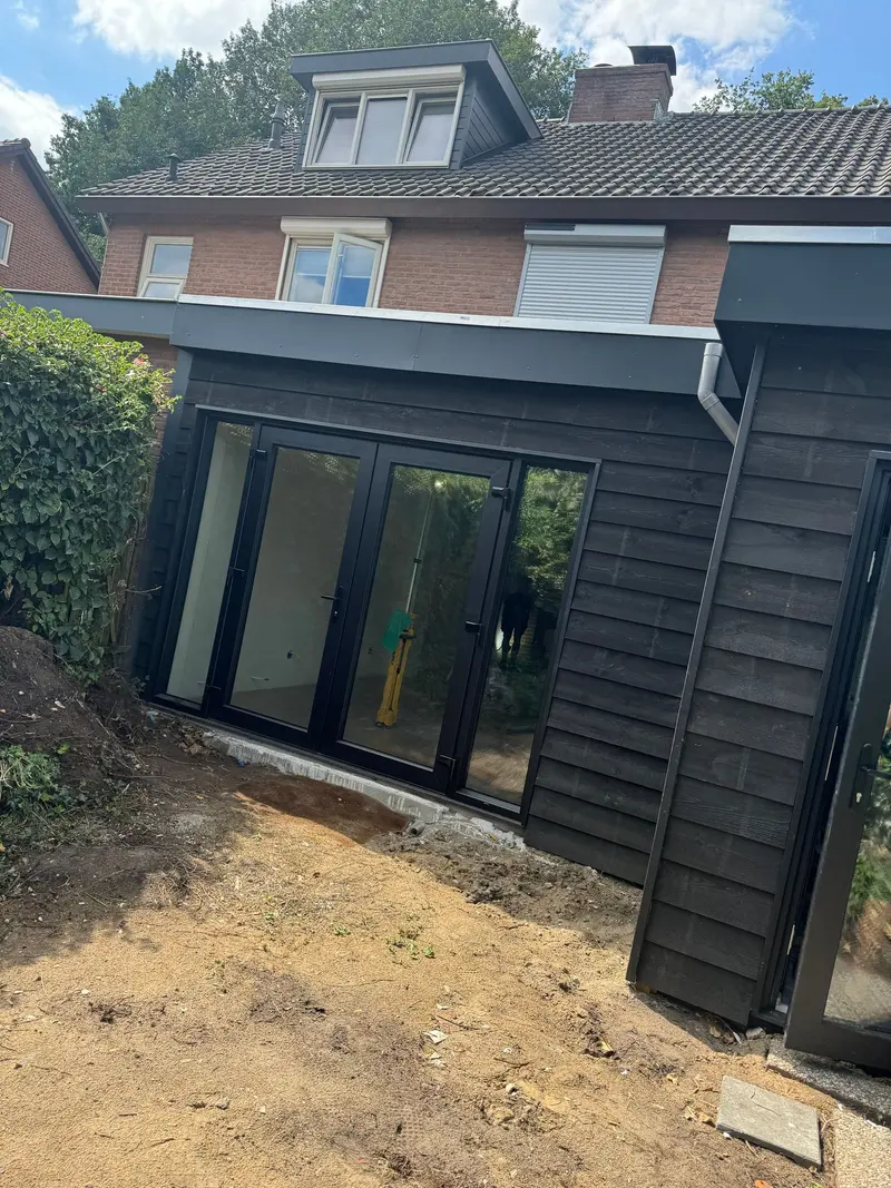 Zijaanzicht van een aanbouw in Breda: zwart gebeitste rabatgevel onder een plat dak, een brede pui met tuindeuren en een aluminium regenpijp op de hoek, tegen een woning met dakkapel