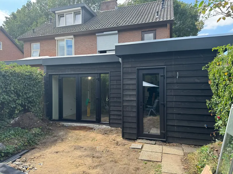 Aanbouw met plat dak aan de achtergevel van een woning in Breda, afgewerkt met zwart gebeitste rabatdelen en een antracietkleurige daktrim, met een pui met tuindeuren en een losse zijdeur; de tuin is nog niet aangelegd