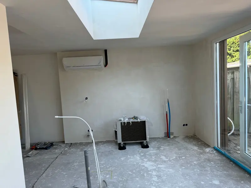 Gestucte ruimte in Etten-Leur met een vierkante lichtkoepel in het plafond en de wandunit van een airco; het buitendeel staat nog ingepakt op de vloer, water- en afvoerleidingen komen uit de wand en de betonvloer is nog niet afgewerkt