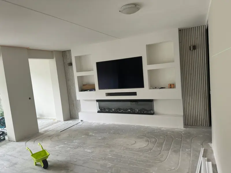 Cinewall in aanbouw in Breda: een gestucte wand met ingebouwde televisie, soundbar en elektrische haard, vier nissen eromheen en verticale houten lamellen aan weerszijden; in de vloer tekenen de lussen van de vloerverwarming zich af
