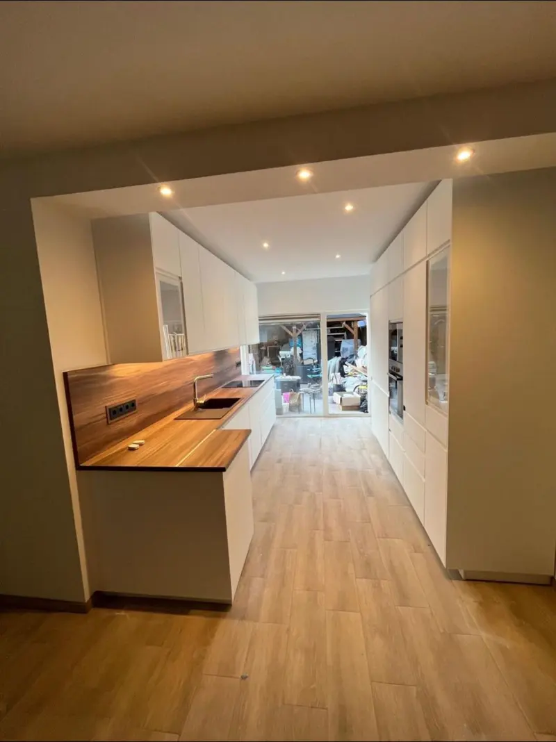 Nieuwe keuken in Capelle aan den IJssel, gezien vanuit de woonkamer door een brede doorgang: greeploze witte kasten met een houten werkblad en bijpassende achterwand, een gouden kraan bij de zwarte spoelbak, een hoge kastenwand met inbouwovens en een houtlookvloer die doorloopt; achter de pui wordt nog gebouwd