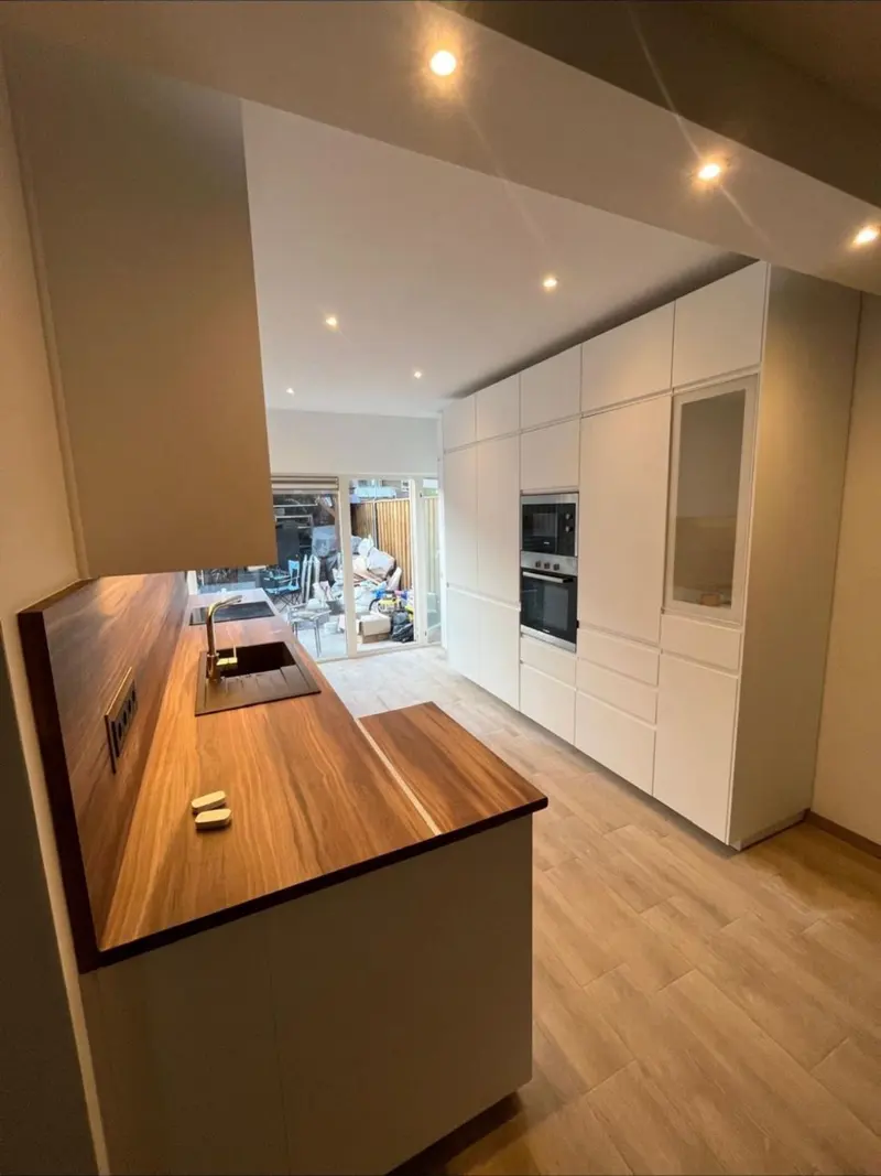 Keuken in Capelle aan den IJssel vanaf het schiereiland: een houten werkblad met gouden kraan, zwarte spoelbak en kookplaat, en daartegenover een hoge witte kastenwand met twee inbouwovens en een kast met matglazen deur