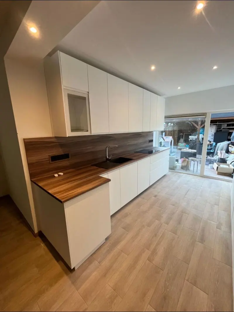 Keukenwand in Capelle aan den IJssel over de volle lengte: witte bovenkasten, een houten achterwand en werkblad met spoelbak en kookplaat, en rechts een schuifpui naar de tuin waar nog bouwmateriaal staat