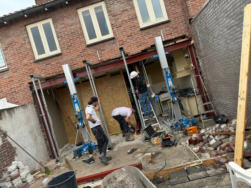 Stalen doorbraak in Rotterdam: in de achtergevel is over de volle breedte een opening gemaakt, afgevangen door een stalen ligger in menie die op stempels rust terwijl het metselwerk erboven wordt gedragen; drie mannen werken aan de ligger, rechts staat de oude keuken nog