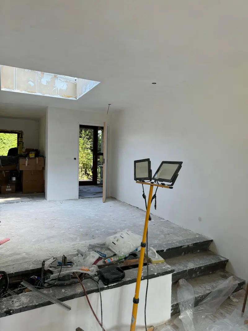 Gestukadoorde en geschilderde woonruimte in Rotterdam, gezien vanaf het lage niveau: de lichtstraat in het plafond, een houten tuindeur naar de tuin en de treden naar het hogere deel; op de betonvloer staan nog bouwlampen en materiaal