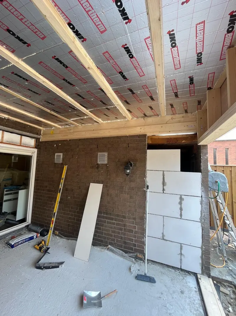 Uitbouw in aanbouw in Breda: het dak is van binnen geïsoleerd tussen de houten balken, de oude buitengevel met ventilatieroosters en buitenlamp staat nu binnen, en een oude opening is dichtgezet met gasbetonblokken