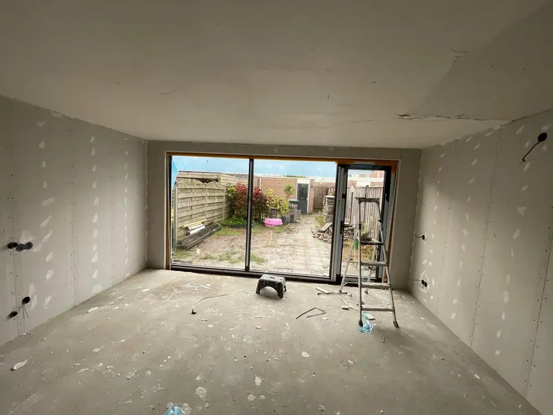 Uitbouw in Breda met de wanden in gipsplaat en de naden gevuld, klaar voor het stucwerk; een zwarte schuifpui met zijdeur kijkt uit op de tuin en de betonvloer is nog kaal
