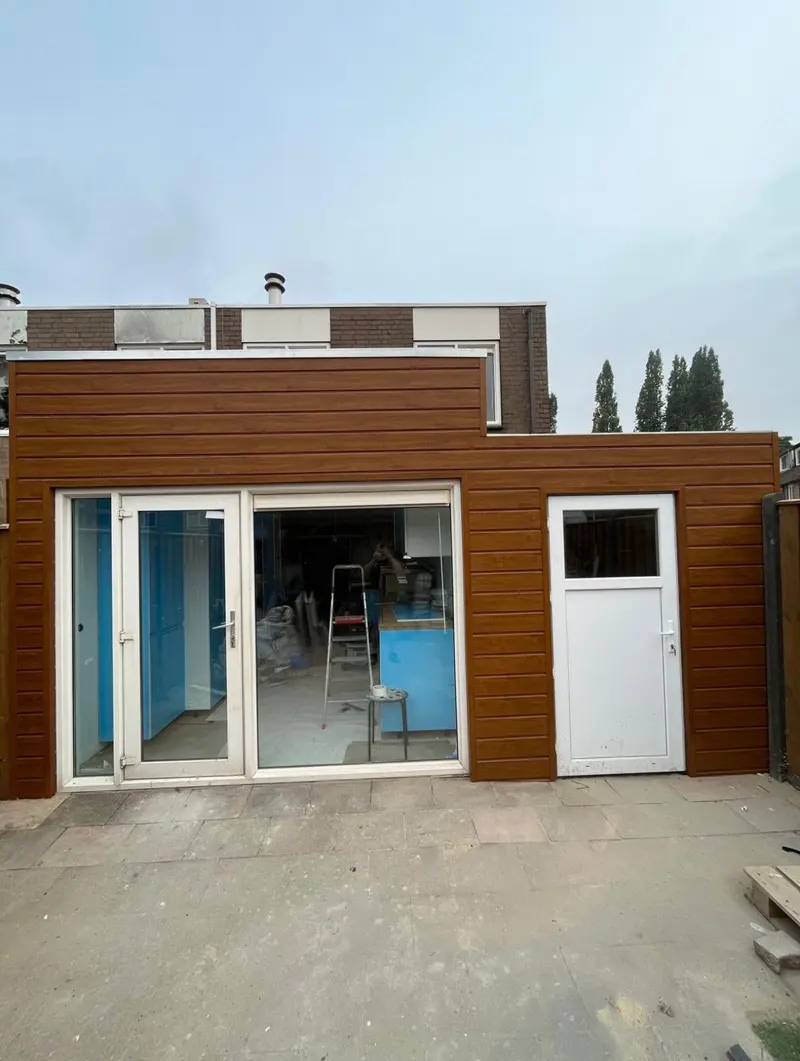 Uitbouw over de volle breedte van de achtergevel van een woning in Breda, afgewerkt met horizontale gevelbekleding in houtlook, met een witte schuifpui, een groot vast raam en een witte achterdeur; binnen ligt nog beschermfolie op het werk