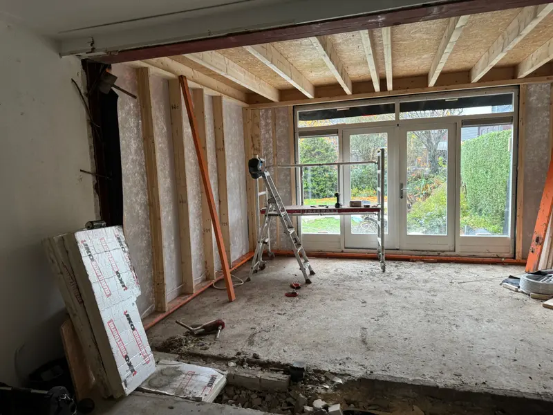 Uitbouw in aanbouw in Breda: houtskeletwanden gevuld met isolatie, een dakconstructie van houten balken met plaatmateriaal erop, en witte openslaande tuindeuren met zijramen; links de doorbraak naar de bestaande woning met een stalen ligger