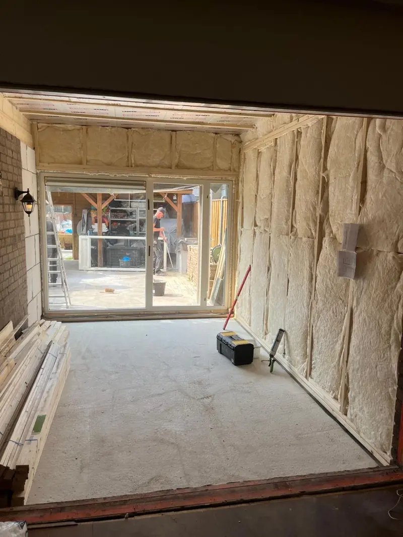 Uitbouw in aanbouw in Breda, gezien vanuit de keuken door de doorbraak: houtskeletwanden gevuld met glaswolisolatie, een schuifpui naar de tuin en een nieuwe dekvloer; op de oude buitenmuur links hangt de buitenlamp er nog