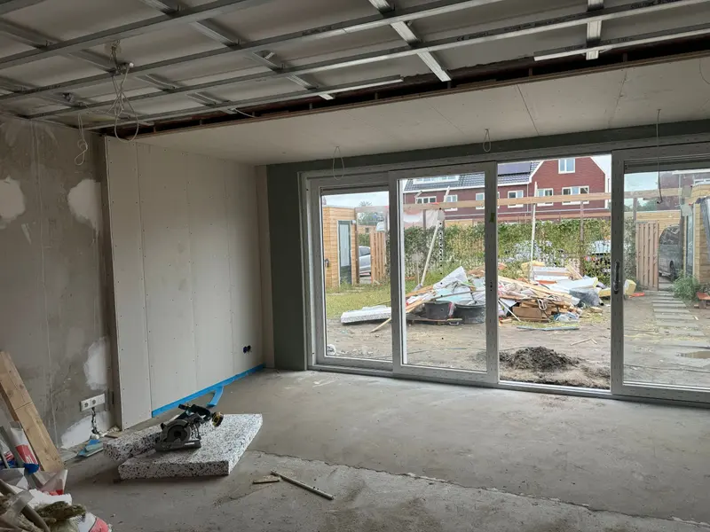 Uitbouw in aanbouw in Breda: stalen plafondprofielen die deels al met gipsplaat zijn dichtgezet, lichtpunten die nog los uit het plafond hangen, een nieuwe binnenwand in gipsplaat en een brede schuifpui naar de tuin; de betonvloer is nog ruw