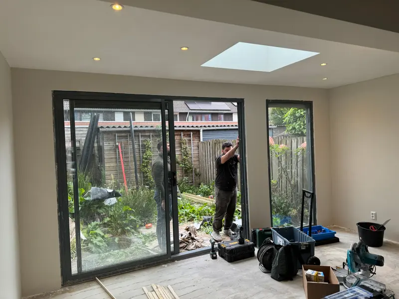 Uitbouw in Breda: gestucte wanden, een plafond met inbouwspots en een lichtkoepel, en een antracietkleurige schuifpui met een hoog zijraam naar de tuin; twee mannen werken aan de pui en de dekvloer is nog kaal