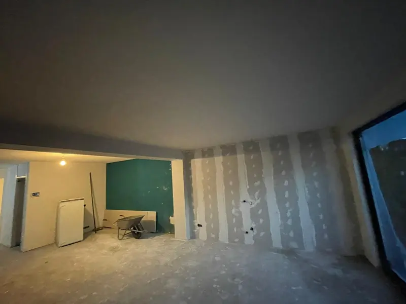 Vanuit de uitbouw in Breda terug de bestaande woning in: rechts de nieuwe gipsplaatwand met gevulde naden, achter de doorgang de oude ruimte met een groen geschilderde wand en een kruiwagen