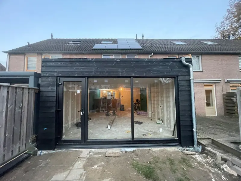 Uitbouw over de volle breedte van de achtergevel van een woning in Breda, bekleed met zwart gebeitste delen, met een brede schuifpui en een zijdeur; door het glas is te zien dat binnen nog gewerkt wordt