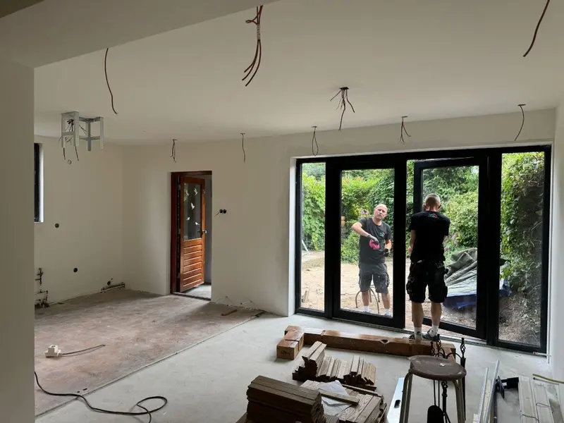 Woning in Rotterdam in de afbouwfase: wanden en plafond zijn gestukadoord en geschilderd, de lichtpunten hangen nog los uit het plafond, en de uitbouw met zwarte pui naar de tuin sluit aan op de bestaande woning; op de betonvloer liggen stapels vloerdelen klaar