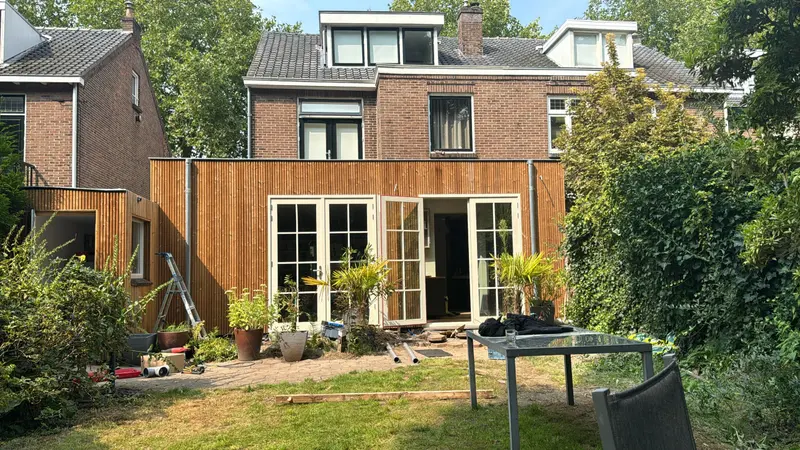 Uitbouw over de volle breedte van de achtergevel van een woning in Rotterdam, bekleed met verticale houten delen, met openslaande tuindeuren met roedeverdeling en verzinkte regenpijpen op de hoeken; in de tuin liggen nog gereedschap en buizen