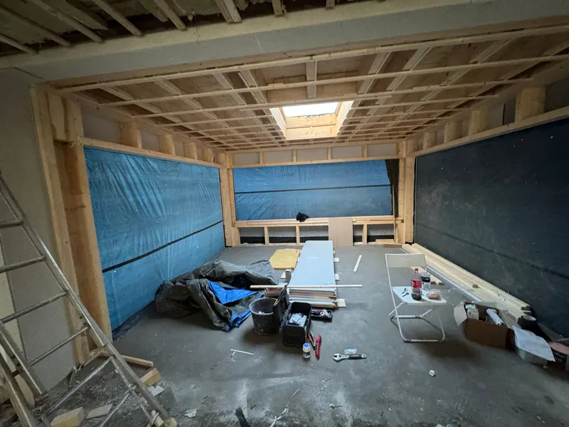 Casco van een uitbouw in Rotterdam: houtskeletwanden en een dakconstructie van houten regels, met een dakraam dat al geplaatst is en wanden die met folie zijn dichtgezet; op de betonvloer liggen gipsplaten, isolatie en gereedschap klaar