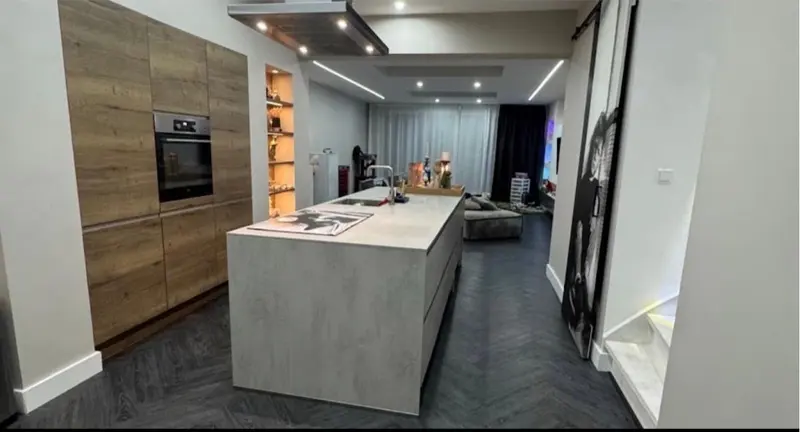 Dezelfde ruimte na de renovatie: een open leefkeuken met een kookeiland in betonlook, een hoge kastenwand in houtlook met inbouwoven, verlichte nissen, een verlaagd plafond met lichtkoven en inbouwspots, en een donkere visgraatvloer die doorloopt naar de woonkamer