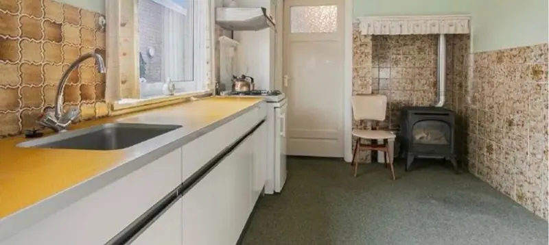 Situatie vóór de renovatie: een smalle, gesloten keuken met een geel werkblad, een roestvrijstalen spoelbak, een gasfornuis onder een afzuigkap en gebloemd tegelwerk tot halverhoogte, met een gaskachel tegen de achterwand en tapijt op de vloer
