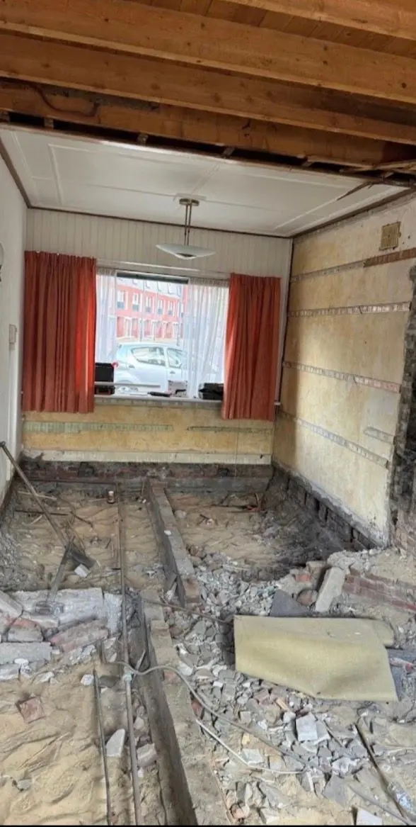 Dezelfde woning tijdens het sloopwerk: de begane grondvloer is volledig uitgebroken tot op het zand, met puin, oude leidingen en de aanzet van het metselwerk in het zicht, en het plafond is open zodat de houten balklaag van de verdiepingsvloer zichtbaar is; op de achtergrond nog de oude schrootjeswand en rode gordijnen