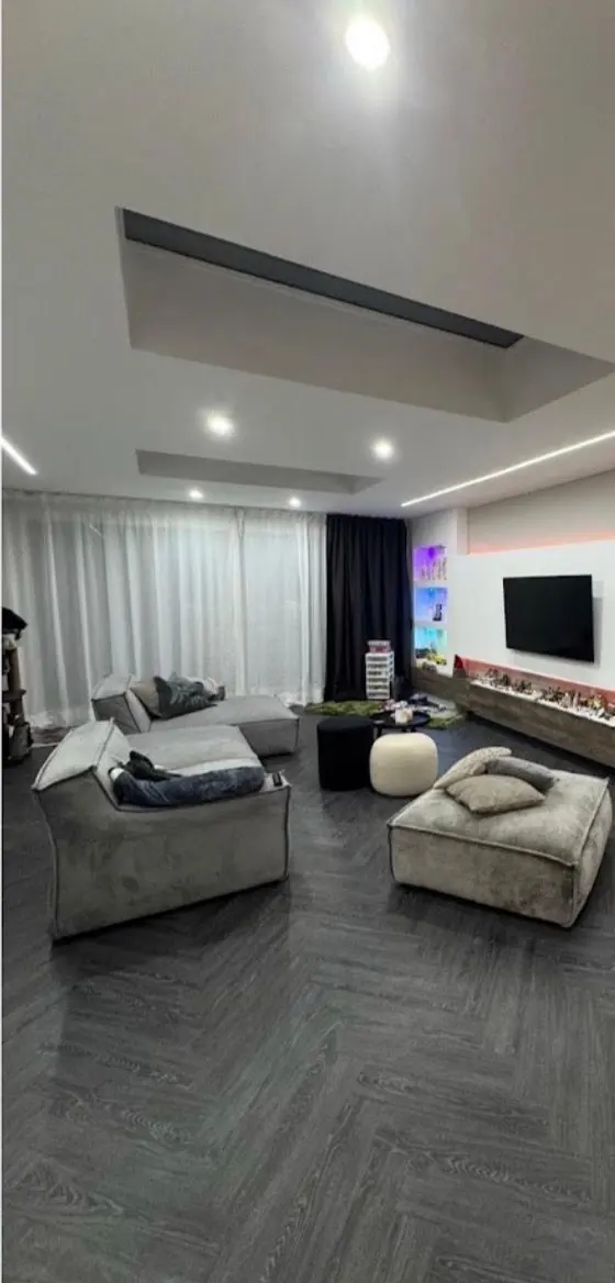 De woonkamer na oplevering: gestucte en geschilderde wanden en plafond met verlaagde koven, indirecte verlichting en inbouwspots, een donkere visgraatvloer, en een wand met tv en verlichte nissen